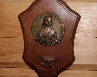 May include: A wooden plaque with a round, embossed metal image of a woman holding flowers. The plaque has a small, decorative metal bowl underneath the image. The text on the image reads "SANCTA TERESIA A JESU INFANTE".
