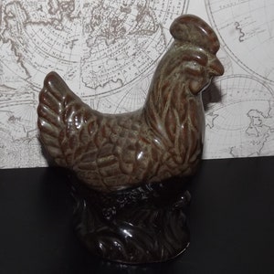 Könnte beinhalten: Eine braune Keramik-Hühnerfigur mit strukturierter Oberfläche. Das Huhn steht auf einem schwarzen Sockel.