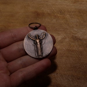 May include: A bronze medal featuring a winged figure holding a wreath. The medal is displayed on a wooden surface.