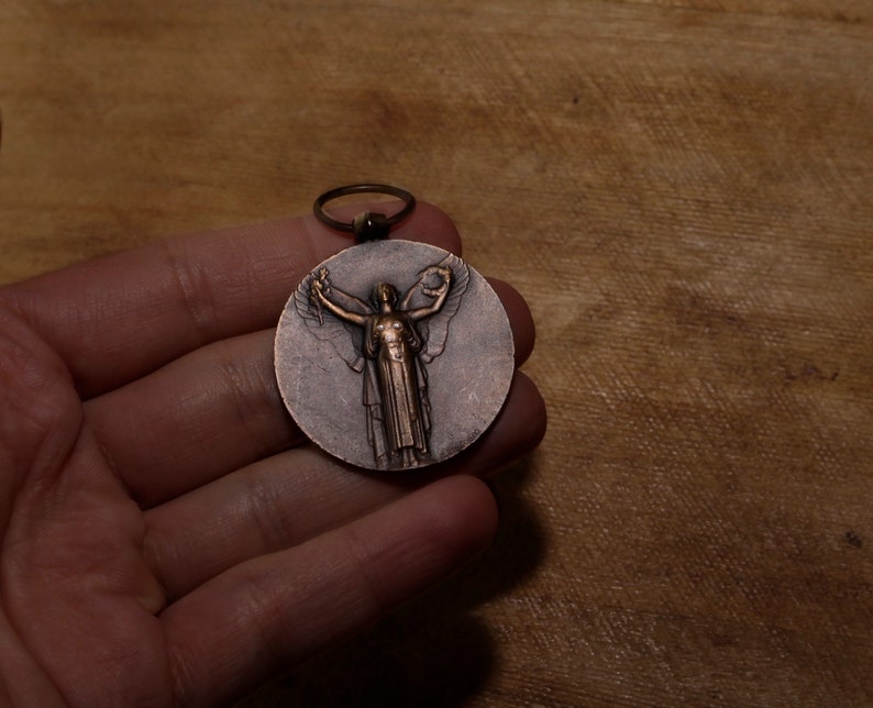 May include: A bronze medal featuring a winged figure holding a wreath. The medal is displayed on a wooden surface.