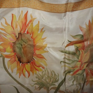 May include: A white silk scarf with a hand-painted design of two sunflowers. The sunflowers are in shades of orange, yellow, and green.