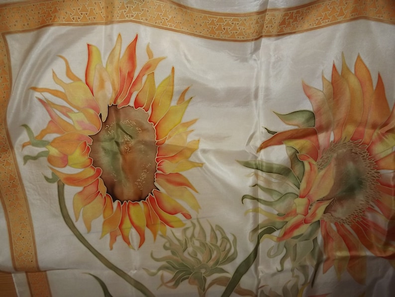 May include: A white silk scarf with a hand-painted design of two sunflowers. The sunflowers are in shades of orange, yellow, and green.