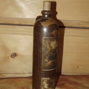 May include: A brown ceramic bottle with a cork stopper and a label that reads "Doppel Kummel" in gold lettering. The bottle has a textured surface and is decorated with gold accents.