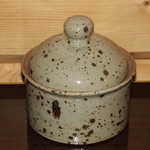 Peut inclure: Un récipient en céramique avec un couvercle, présentant un glaçage moucheté brun et blanc. Le récipient a une forme arrondie et une finition lisse et brillante.