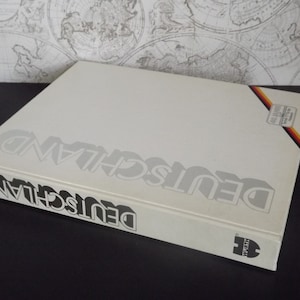 Puede incluir: Un libro blanco con el título "DEUTSCHLAND" impreso en letras negras y grises. El libro tiene una franja negra y amarilla en la portada con un pequeño logotipo en la esquina inferior derecha.