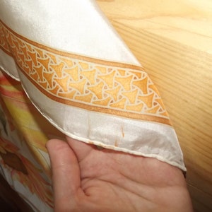 May include: A white silk scarf with a gold and white geometric pattern border. The scarf has a floral design in shades of orange and yellow.