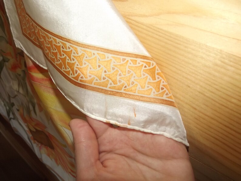 May include: A white silk scarf with a gold and white geometric pattern border. The scarf has a floral design in shades of orange and yellow.