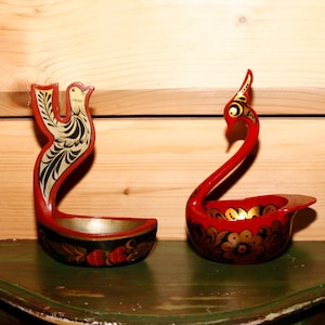 Op de afbeelding: Twee handgeschilderde houten opscheplepels. De ene is een rode lepel met een vogelontwerp, de andere is een rode zwaanvormige lepel. Beide lepels hebben gouden en zwarte bloemaccenten en worden tentoongesteld op een groen oppervlak.