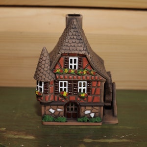 Può includere: Una piccola casa in ceramica marrone con una facciata in mattoni rossi e un tetto marrone. La casa ha una piccola finestra sul davanti e una piccola porta. C'è una piccola ruota sul lato della casa.