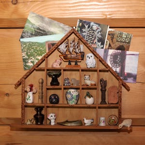 Peut inclure: Une étagère en bois en forme de maison remplie de figurines miniatures et de cartes postales. L'étagère contient un navire, un hibou, un chat, un cochon et d'autres petits objets décoratifs. Le fond est un mur en bois.