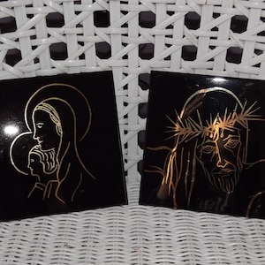 May include: Two black square tiles with gold outlines of religious figures. The left tile depicts the Virgin Mary holding a baby Jesus. The right tile depicts Jesus with a crown of thorns.