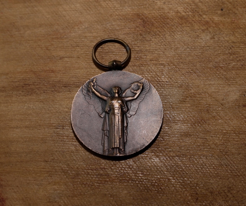 May include: A bronze medal featuring a raised image of a woman holding a wreath in her outstretched arms. The woman is wearing a flowing robe and has her hair pulled back. The medal is suspended from a ring.