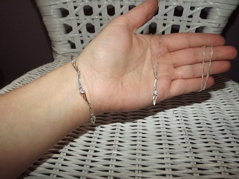 Peut inclure: Un collier en argent avec un pendentif d&eacute;licat pr&eacute;sentant un motif stylis&eacute; avec de petites pierres claires. Un bracelet en argent assorti avec un design similaire est &eacute;galement pr&eacute;sent&eacute;.