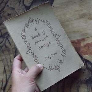 May include: A vintage book titled "A Book of French Songs" by St&eacute;phan. The book cover is beige with a decorative black border.