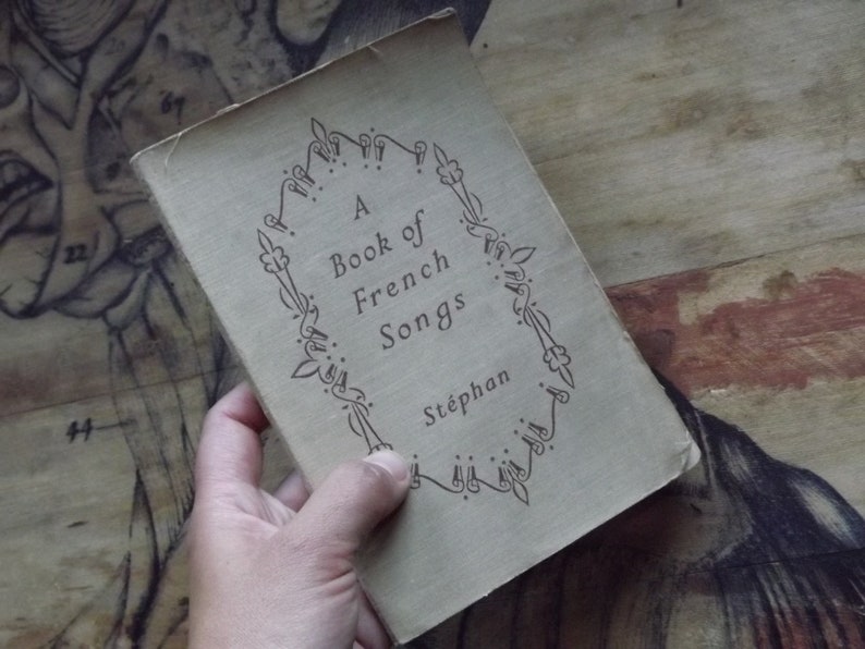 May include: A vintage book titled "A Book of French Songs" by St&eacute;phan. The book cover is beige with a decorative black border.