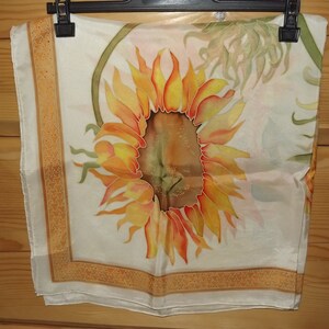 May include: A white silk scarf with a hand-painted sunflower design. The sunflower is yellow, orange, and brown with green leaves. The scarf has a gold border with a geometric pattern.