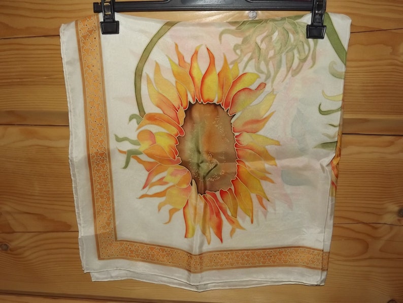 May include: A white silk scarf with a hand-painted sunflower design. The sunflower is yellow, orange, and brown with green leaves. The scarf has a gold border with a geometric pattern.