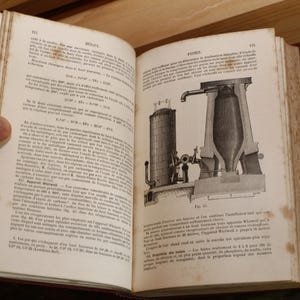 Può includere: Un libro antico aperto con pagine ingiallite e invecchiate. La pagina sinistra presenta testo, mentre la pagina destra mostra una dettagliata illustrazione in bianco e nero di macchinari industriali. La copertina del libro è rosso scuro.