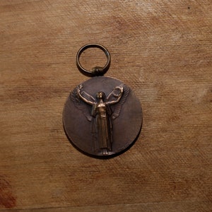 May include: A bronze medal with a ring for hanging. The medal features a raised image of a woman with wings outstretched, holding a wreath in her right hand.