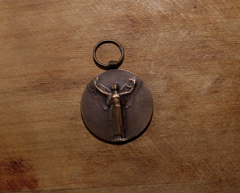 May include: A bronze medal with a ring for hanging. The medal features a raised image of a woman with wings outstretched, holding a wreath in her right hand.
