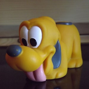 Könnte beinhalten: Ein gelber Spielzeug-Hund aus Gummi mit schwarzen Ohren und einer herausgestreckten rosa Zunge. Der Hund liegt auf der Seite mit dem Kopf nach links gedreht.