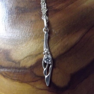 Peut inclure: Un collier avec un pendentif en argent, au design abstrait et d&eacute;licat, orn&eacute; d'une seule pierre transparente. La cha&icirc;ne est une double cha&icirc;ne en argent.