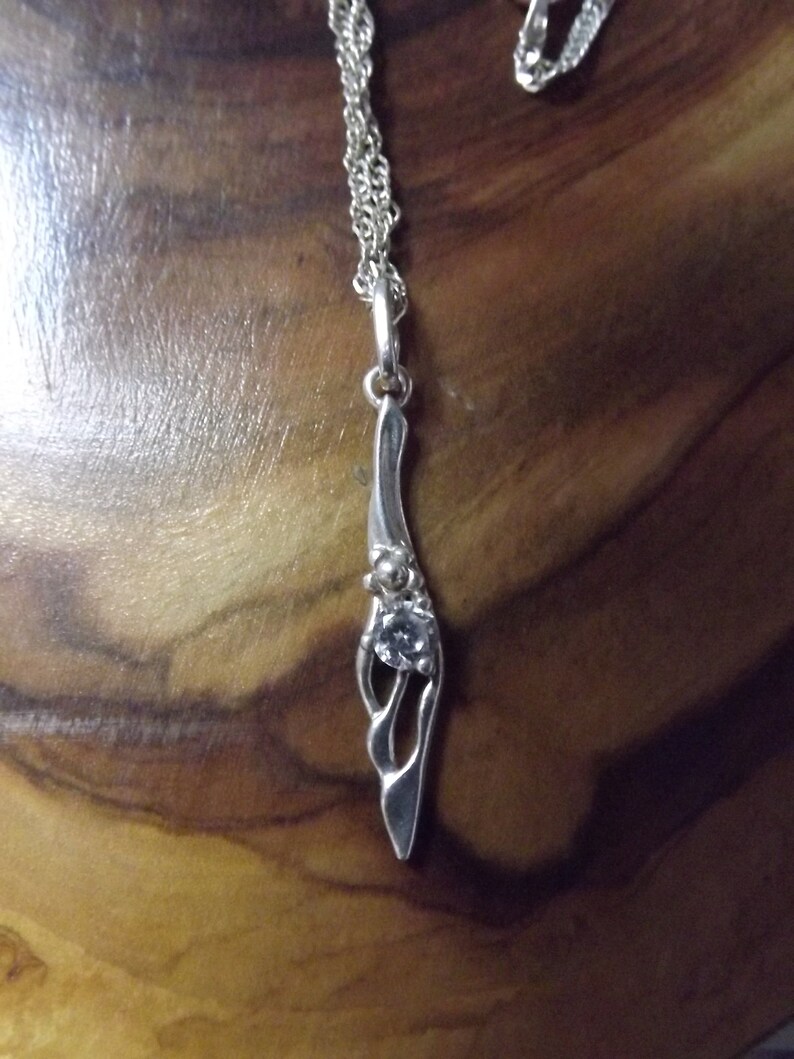 Peut inclure: Un collier avec un pendentif en argent, au design abstrait et d&eacute;licat, orn&eacute; d'une seule pierre transparente. La cha&icirc;ne est une double cha&icirc;ne en argent.