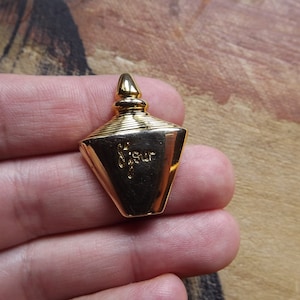 Puede incluir: Un colgante dorado con forma de frasco de perfume. El colgante tiene una parte superior puntiaguda y una base con la palabra "fleur" inscrita. El frasco se sostiene en una mano.