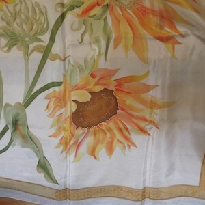 May include: A white silk scarf with a hand-painted design of sunflowers and green leaves. The scarf has a gold border.