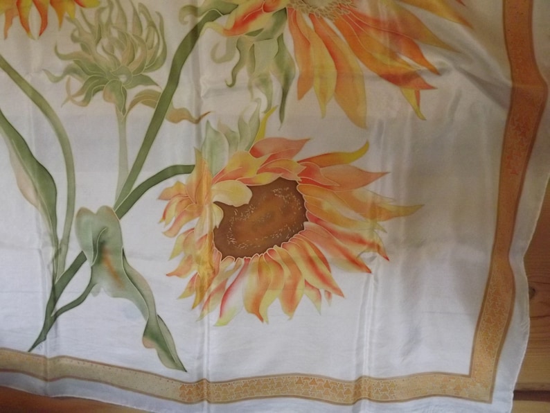 May include: A white silk scarf with a hand-painted design of sunflowers and green leaves. The scarf has a gold border.