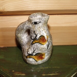 May include: A speckled grey ceramic jug with a handle and a spout. The jug is decorated with two deer figures in orange and brown, with black antlers. A black eye is painted on the side.