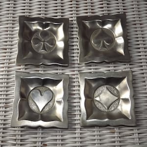 May include: Four silver-colored metal trinket dishes with raised designs of playing card suits: clubs, spades, hearts, and diamonds.
