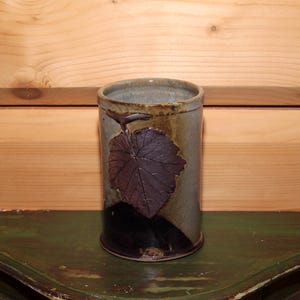 Peut inclure: Un vase cylindrique en céramique avec un motif de feuille. Le vase est principalement gris-vert avec une feuille et une tige brun foncé. Le bas du vase est brun foncé et noir. Le vase est posé sur une surface verte.