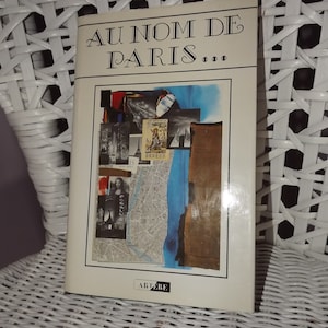 May include: A white book with the title "AU NOM DE PARIS..." in black text. The cover features a collage of images, including a map of Paris, a blue sky, and a building.
