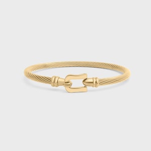 Może przedstawiać: Złota bransoletka ze skręconym kablem. Bransoletka posiada zapięcie w kształcie litery U pośrodku. Biżuteria jest prezentowana na jasnoszarym tle. Obwód bransoletki wynosi około 18 cm.