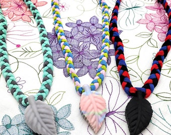 May include: Three braided fabric necklaces with leaf-shaped pendants. The necklaces are in different colors: teal and gray, blue, green, and pink, and red and black. The leaf pendants are gray, pink and white, and black.