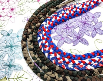 May include: Braided rope necklaces in various colors, including camouflage, brown, and red, white, and blue. The necklaces are arranged on a floral patterned fabric, showcasing their textures and designs.