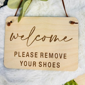以下が含まれることがあります： 上部に茶色の革紐がついた木製の看板。看板には「ようこそ」と筆記体で、「靴を脱いでください」とブロック体で書かれています。