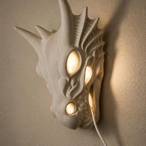 May include: A white, dragon-shaped wall sconce with three illuminated eye-shaped lights. The sconce is mounted on a textured, light-colored wall. A white power cord extends from the sconce to a wall outlet.