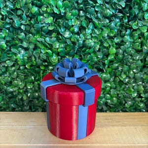 May include: A red cylindrical gift box with blue vertical stripes and a blue bow on top. The box is sitting on a wooden surface with a green leafy background.