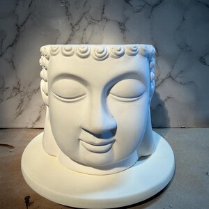 May include: A white Buddha head planter with closed eyes and a serene expression. The planter is on a round base and features detailed hair and facial features. The neutral color and design make it suitable for various decor styles.
