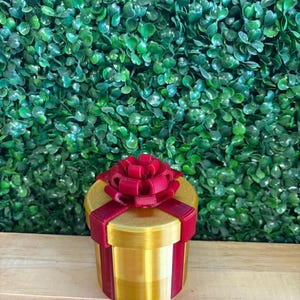 May include: A gold-colored, cylindrical gift box with a red bow on top and red ribbon accents. The box is sitting on a wooden surface in front of a green leafy background. The gift box is a decorative item.