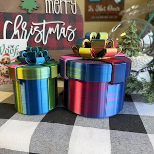 May include: Two colorful, cylindrical gift boxes with decorative bows. One is blue and green, the other is red and blue. The background includes a sign that says "Merry Christmas". The boxes are on a black and white checkered surface.