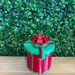 May include: A 3D-printed, round gift box in red and green, topped with a red bow. The box has a cylindrical shape with a green lid and a red body. The bow is intricately designed, adding a festive touch. The background is a wall of green foliage.