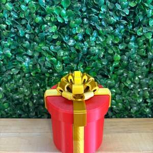 May include: A red, cylindrical gift box with a gold bow and ribbon. The box is sitting on a wooden surface in front of a green leafy background. The bow is large and ornate, with multiple loops and a textured appearance.