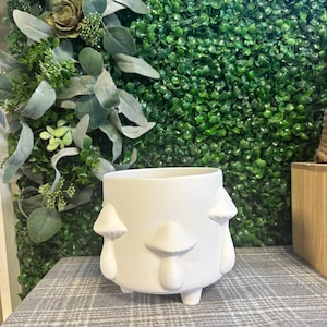 May include: A white ceramic planter with mushroom-shaped embellishments. The planter has a round shape with three small feet and is set against a backdrop of green foliage. The mushrooms are evenly spaced around the planter.