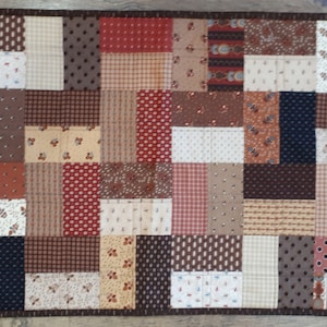May include: A patchwork quilt runner featuring a mix of brown, beige and red fabrics. The runner is composed of squares and rectangles of varying sizes and patterns.
