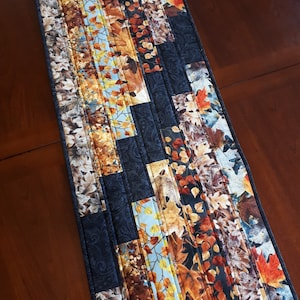Peut inclure: Un chemin de table matelassé avec un motif de feuilles d'automne dans des tons de brun, orange, jaune et bleu. Le chemin de table est fait de tissu avec un fond noir et un motif cachemire.