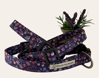 Puede incluir: Dos collares para perros con un patrón floral en tonos de morado, rosa y blanco sobre un fondo azul oscuro. Los collares tienen hebillas negras y un anillo en D plateado. Los collares están etiquetados como "Blonde Dog Designs".