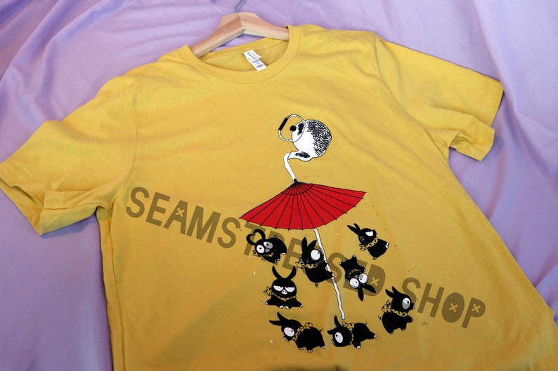 May include: Mustard yellow t-shirt with a whimsical illustration. The design features a figure with a white head and red umbrella, surrounded by black bunny-like creatures. The text "SEAMS SHOP" is printed on the shirt.